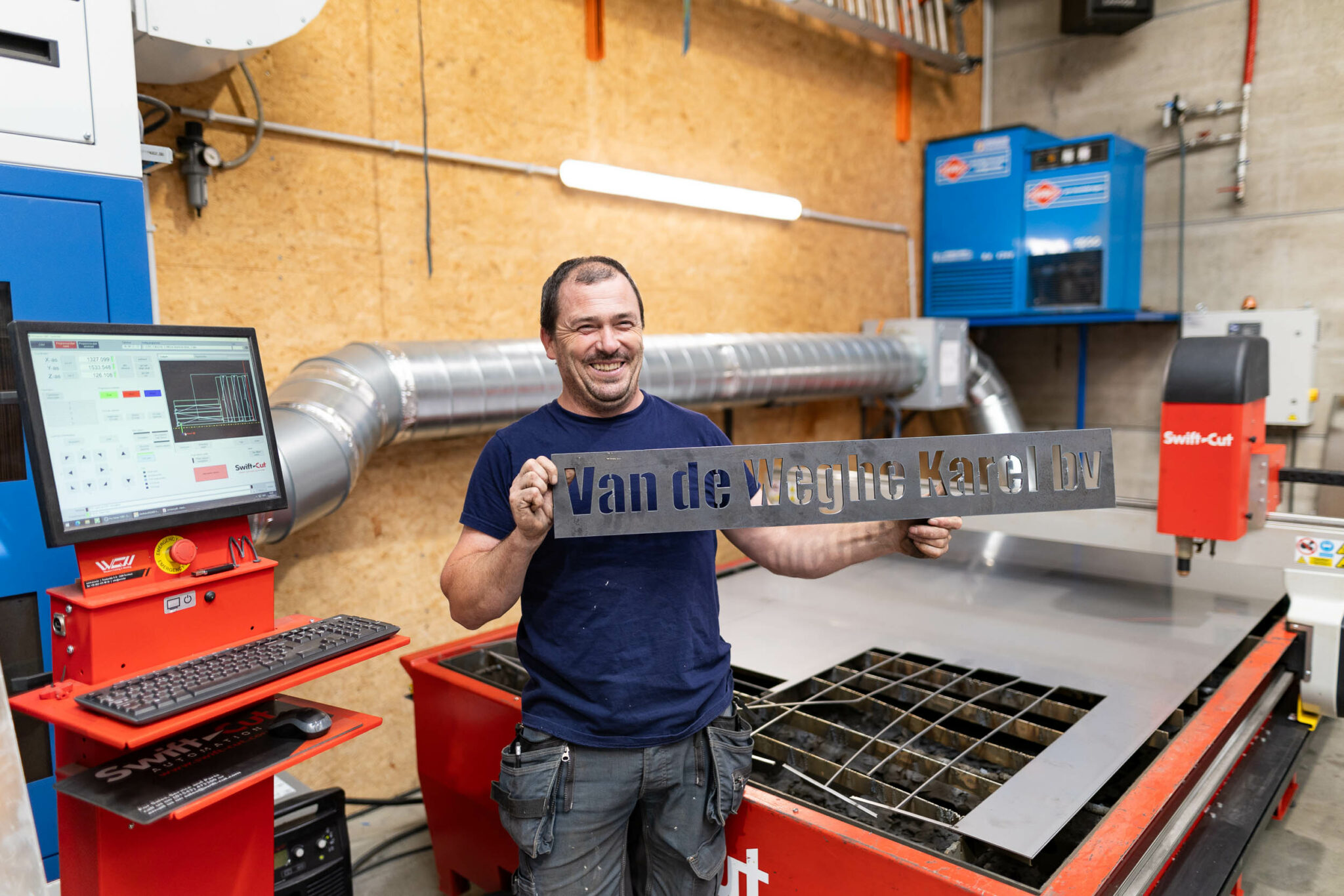 Een medewerker van Van de Weghe Karel BV staat voor een Swift-Cut Pro CNC-plasmasnijtafel met zijn naam, uitgesneden uit plaatstaal, in zijn handen
